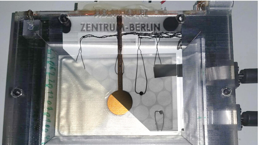 Fotomontage: Die Diagonale teilt das Bild in ein Foto der Lithium-Schwefel-Pouchzelle (links) und das zugeh&ouml;rige R&ouml;ntgenbild (rechts) w&auml;hrend der multimodalen Messung mit Kraftsensor (golden) sowie Temperatursensoren. Auf dem R&ouml;ntgenbild ist die perforierte Wabenstruktur des Stromkollektors gut zu erkennen. Dieser neue Designansatz reduziert das Gewicht der Zelle, ohne die Leistung zu beeintr&auml;chtigen.